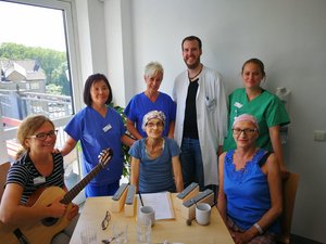 Bild zeigt Mitarbeiterin mit Gitarre, zwei Patientinnen und weitere Team-Mitglieder